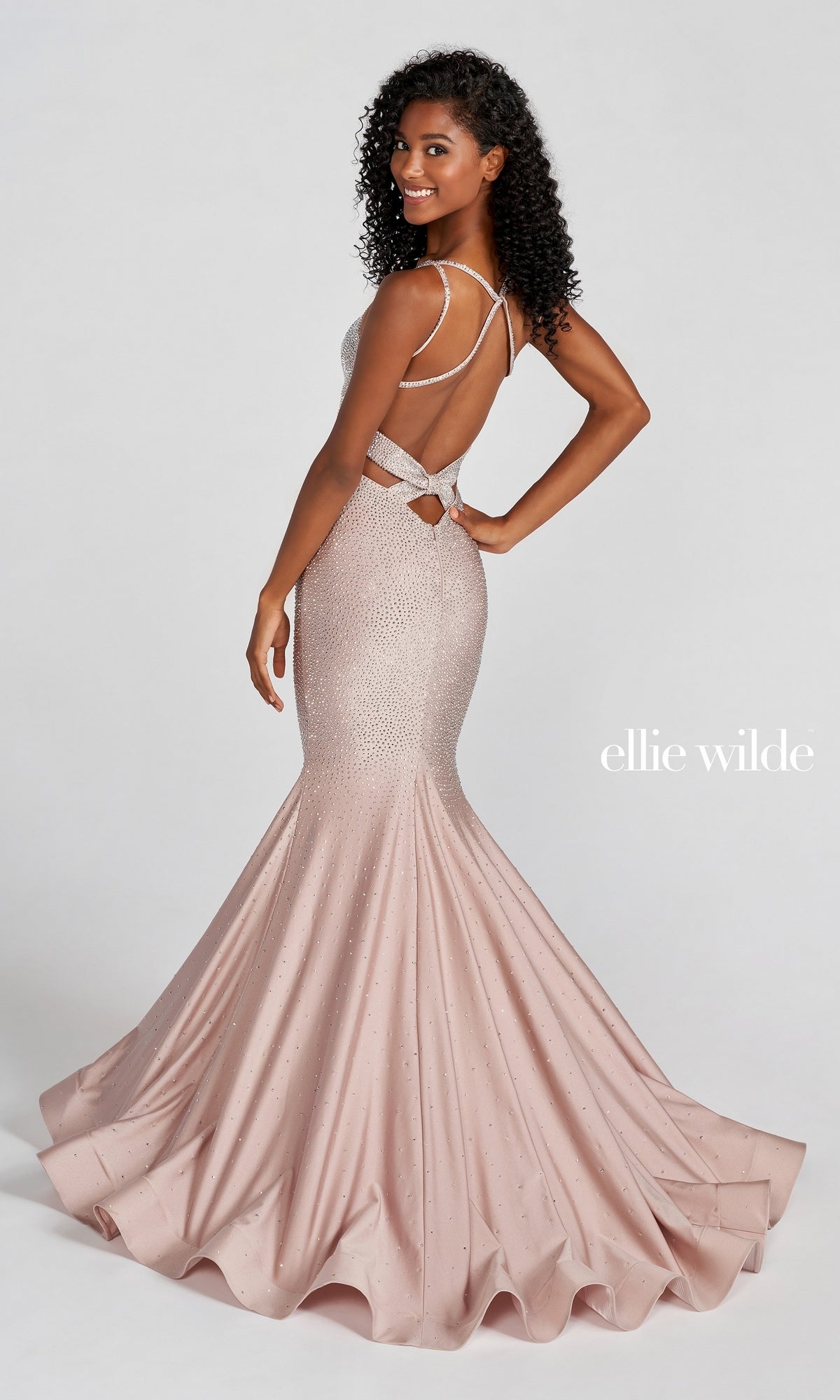 A woman poses, smiling over her shoulder in the Strappy-Back Long Beaded Ellie Wilde Formal Gown—a blush pink mermaid dress with a flared bottom, open back, crisscross straps, and elegant beaded embellishments.